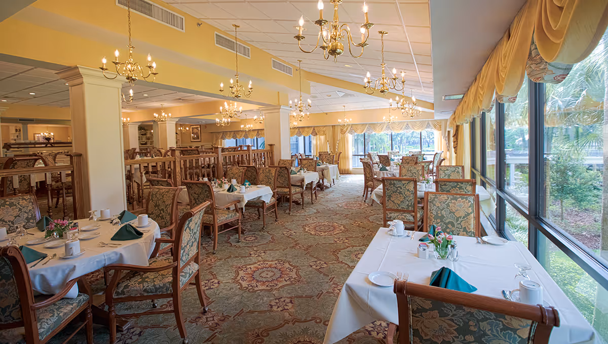 Bright, elegant dining room with multiple white-clothed tables and upholstered chairs, chandeliers overhead and large windows along one side.