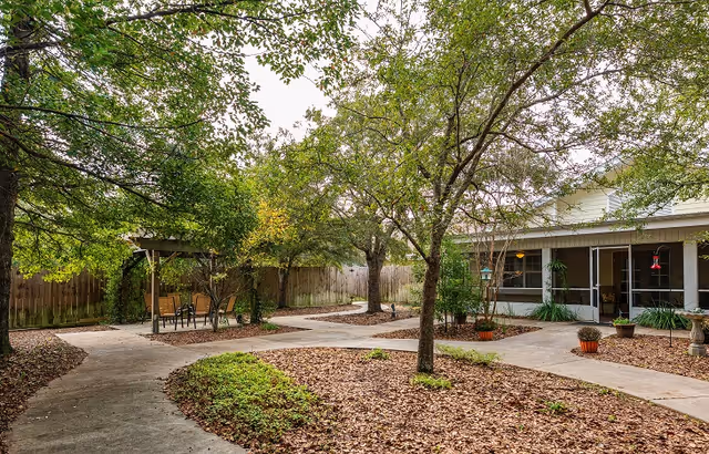 Outdoor courtyard area with paved walkways, trees, and seating under a small pergola. The courtyard is surrounded by a wooden fence and adjacent to a building with large windows and potted plants.