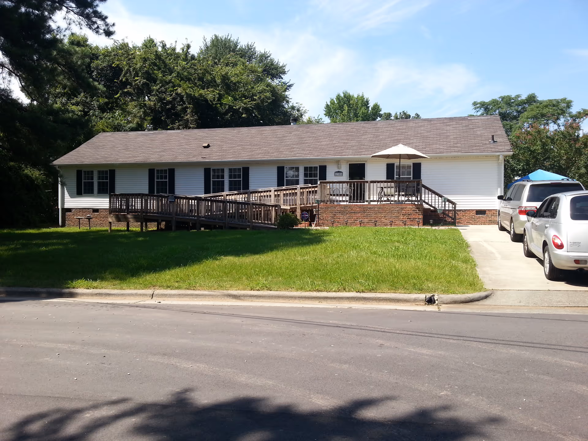 Single-story white building with black shutters and a brown shingled roof. There is a wooden wheelchair ramp and a small porch with a table and umbrella in front. Two cars are parked on the driveway to the right. The building is surrounded by green grass and trees under a blue sky.