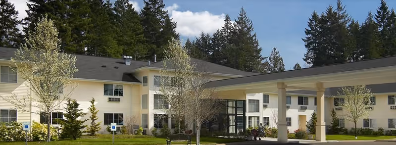 Front exterior of a two-story light-colored assisted living building with a covered drop-off entrance, lawn, trees, and surrounding evergreens.