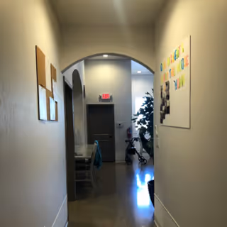 A well-lit hallway in a senior living facility with bulletin boards on the walls, an archway to a communal area, and a walker near a doorway.