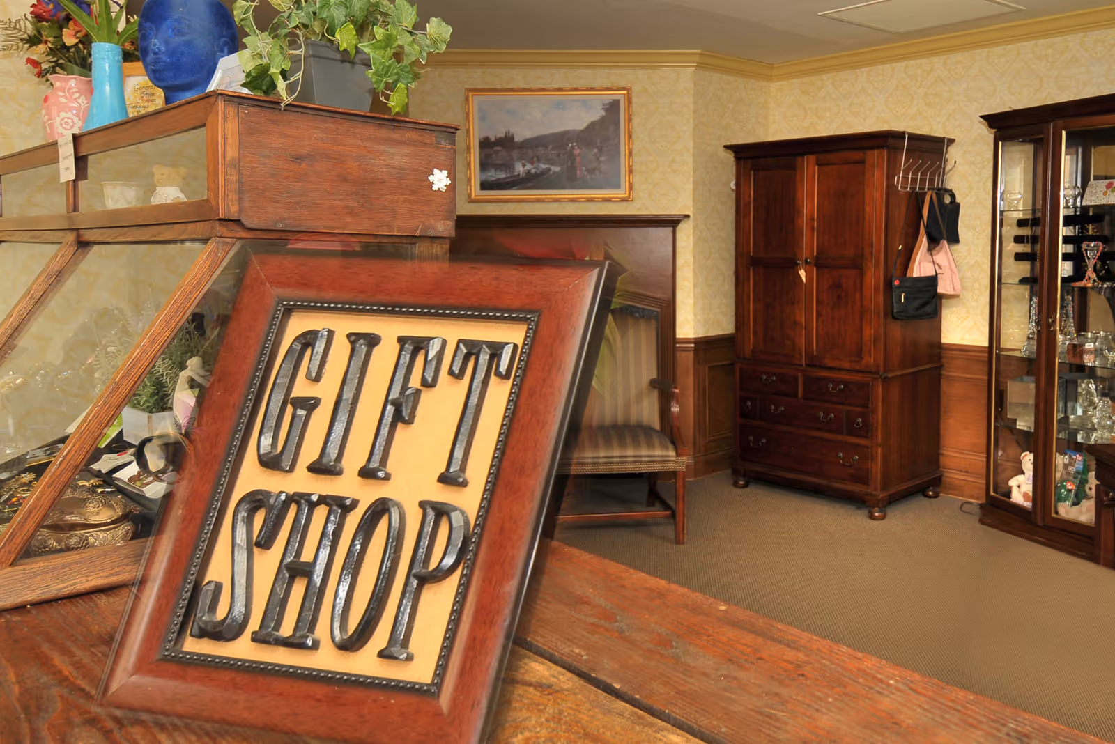Framed 'GIFT SHOP' sign on a wooden counter in an interior gift shop with display cabinets and furniture.