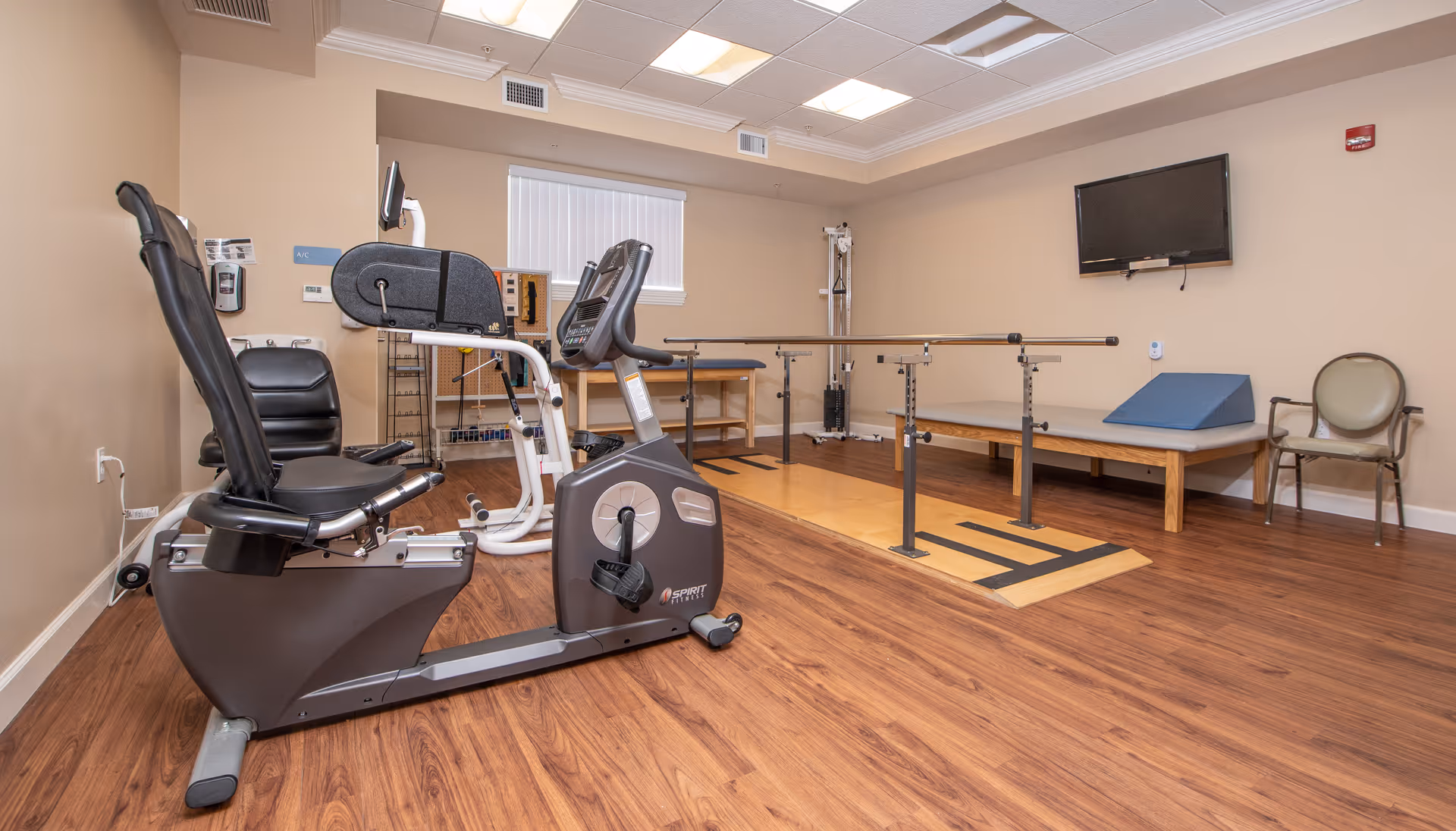 A rehabilitation or physical therapy room with exercise equipment including a recumbent bike, parallel bars for walking support, a therapy table with a blue wedge cushion, a wall-mounted TV, and a chair against the beige walls and wood flooring.