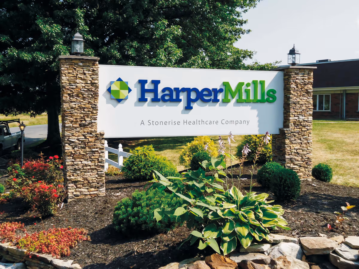 Outdoor view of a healthcare facility sign for Harper Mills, a Stonerise Healthcare Company, mounted between two stone pillars with landscaping including bushes and flowers in front.