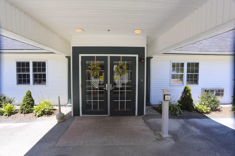Covered entrance to a building with double glass doors decorated with hanging flower baskets. The building exterior is white with black trim around the doors and windows. There are small landscaped areas with shrubs and plants on either side of the entrance.