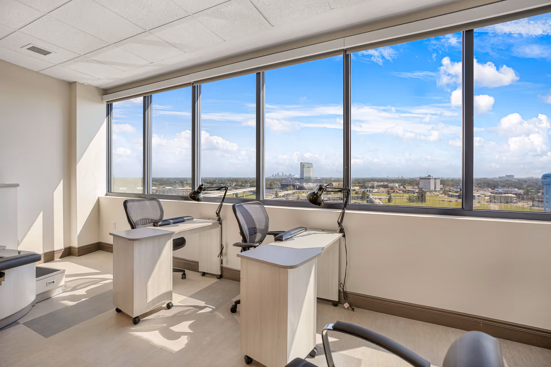 Sunlit interior office-like room with two desks and chairs facing large windows overlooking a city skyline.