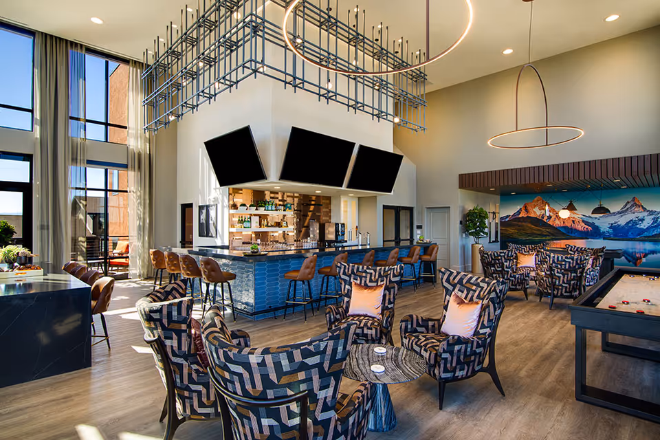 A spacious and modern common area with patterned armchairs and small round tables arranged for seating. There is a bar with high stools and three large black screens mounted above it. The room features large windows with sheer curtains, contemporary circular light fixtures, and a mural of mountains on one wall. A shuffleboard table is visible on the right side of the image.