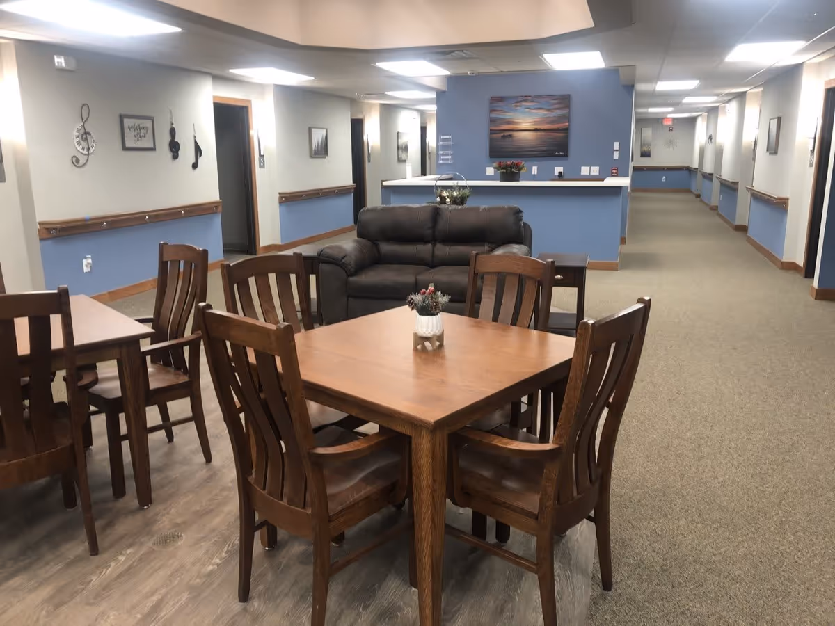 Interior view of a senior living facility common area with wooden tables and chairs, a dark brown leather loveseat, and a blue accent wall with a landscape painting. The hallway extends on both sides with doors and wall decorations including musical notes and framed pictures.