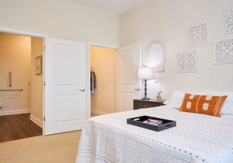 Light, neutral bedroom with a made bed, bedside table and lamp, and open doors leading to a bathroom and closet.