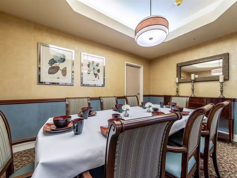 A formal dining room with a long table covered with a white tablecloth, set with red plates, bowls, and black mugs. The room has beige and blue walls, two framed botanical prints, a large mirror above a wooden sideboard with three candlesticks, and a ceiling light fixture.