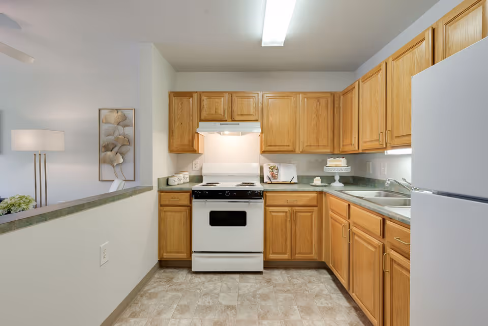 A clean kitchen with wooden cabinets, a white stove with an overhead vent, a white refrigerator, and a double sink. The kitchen has green countertops and a tiled floor. To the left, there is a partial view of a living area with a floor lamp and wall art featuring metallic leaves.