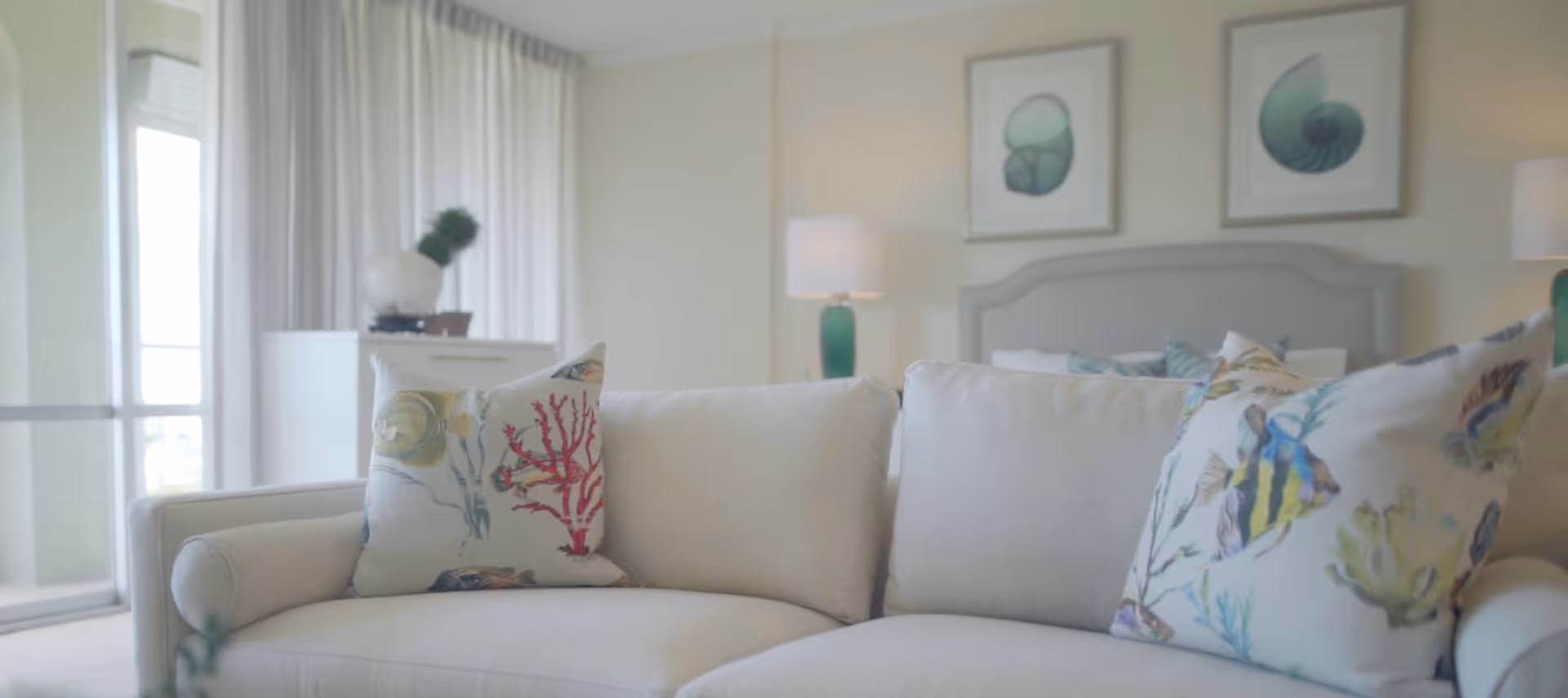Light-filled bedroom suite with a cream sofa and decorative pillows in the foreground and a bed with lamps and framed art in the background.