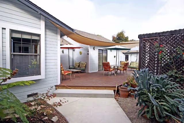 Outdoor patio area at Villas Assisted Living with wooden deck, tables, chairs, umbrellas, and surrounding plants and shrubs. The patio is adjacent to light gray buildings with windows and a lattice fence on the right side.