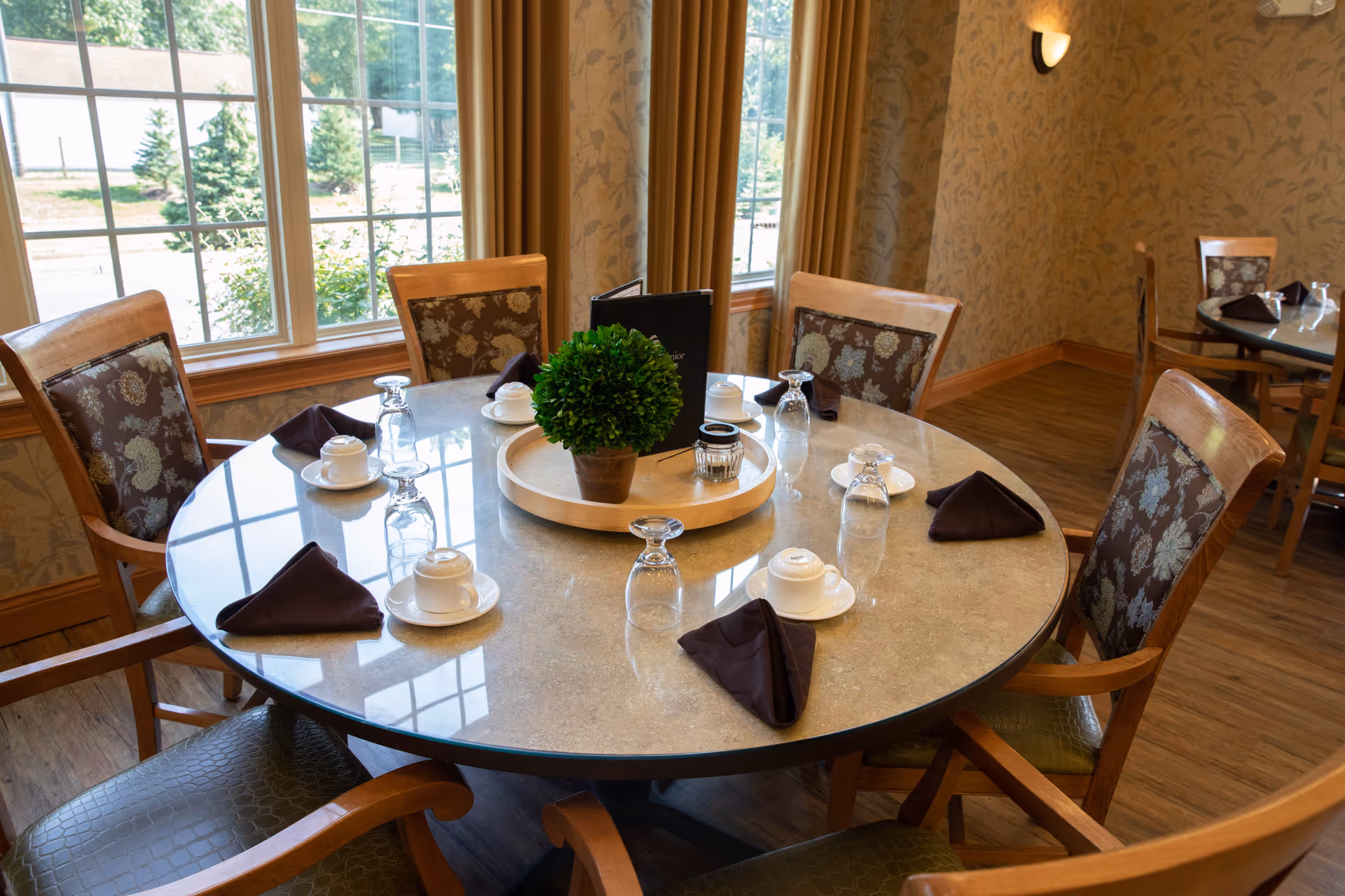 A round dining table set for six with wooden chairs featuring patterned cushions. The table has a centerpiece with a small green plant in a pot on a wooden tray, along with upside-down glasses, cups on saucers, and folded dark napkins. The room has large windows with beige curtains and floral wallpaper, and wooden flooring.