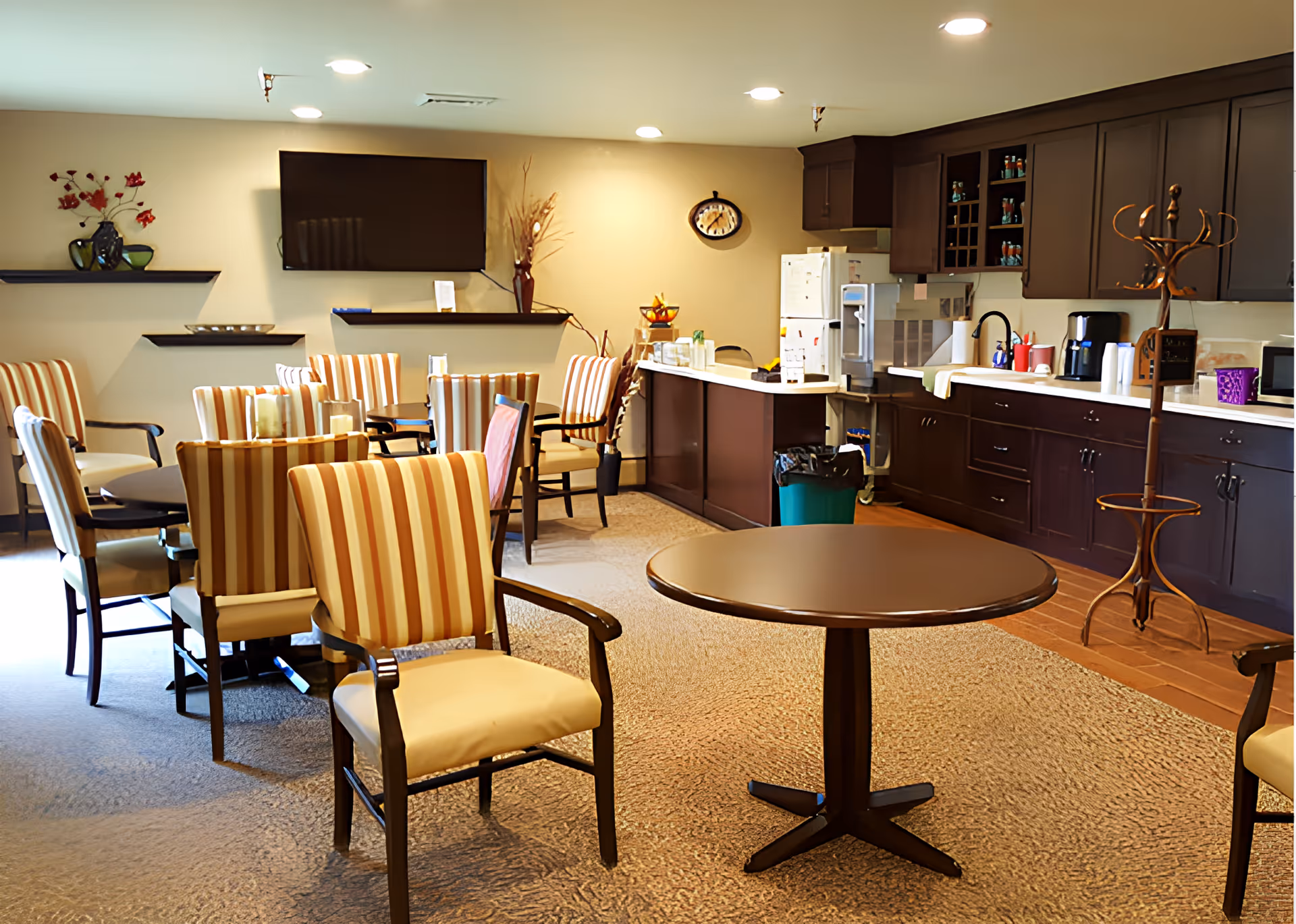 A cozy common area with multiple striped upholstered chairs arranged around small tables. The room features a wall-mounted flat-screen TV, decorative shelves with vases and flowers, a clock on the wall, and a kitchenette with dark wood cabinets, a refrigerator, coffee maker, and other kitchen appliances. There is also a coat rack near the kitchenette.