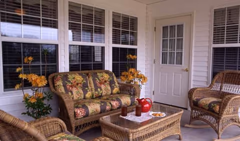 A cozy enclosed porch area with wicker furniture including a sofa, two chairs, and a coffee table. The sofa and chairs have floral patterned cushions. There are potted yellow flowers on either side of the sofa. On the coffee table, there is a red teapot, two glasses, and a plate with cookies. The porch has large windows with white frames and a white door with a window panel.