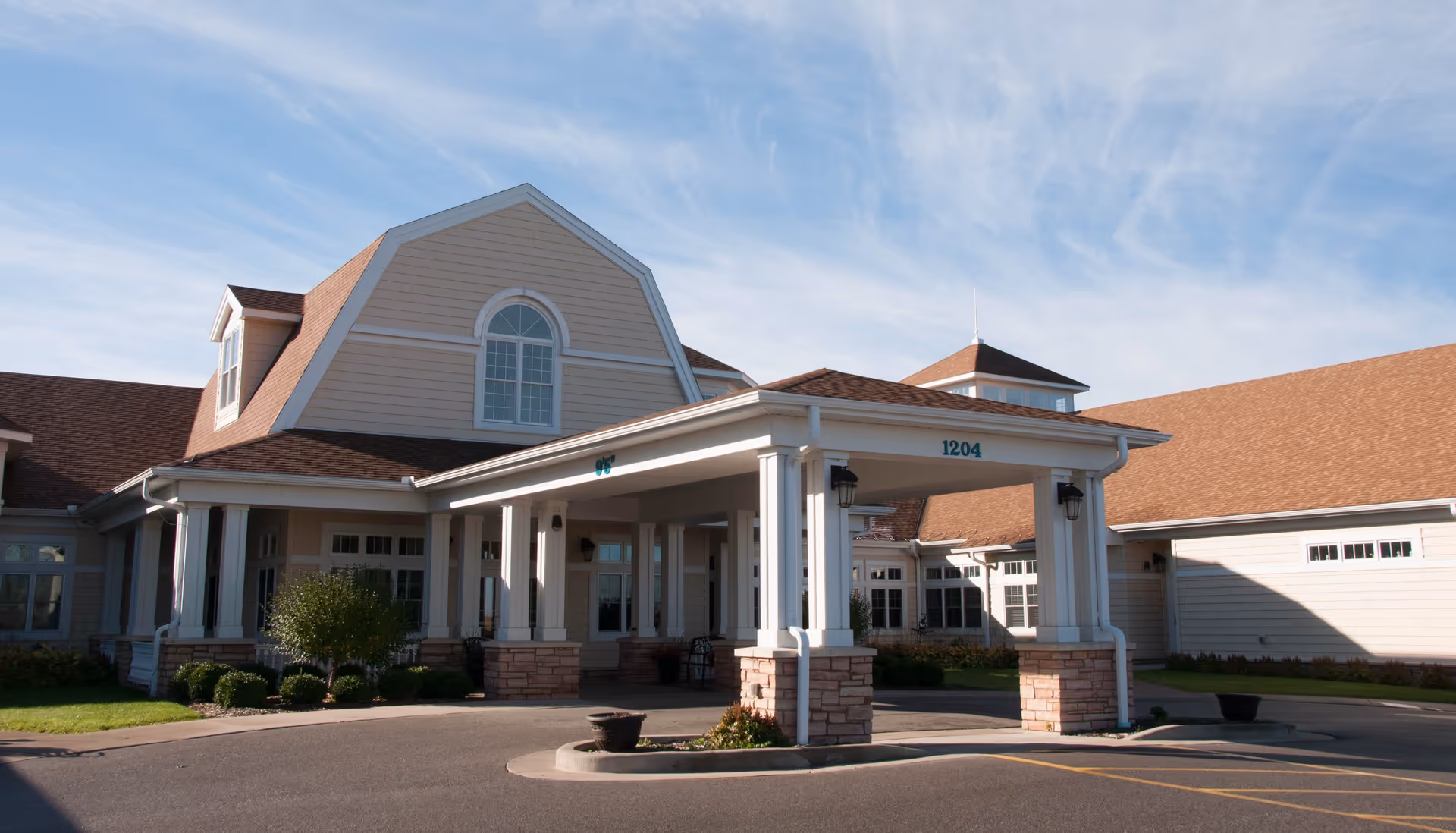 Exterior front view of a senior living facility building with a covered entrance supported by white columns and stone bases, a driveway in front, and a clear blue sky above.