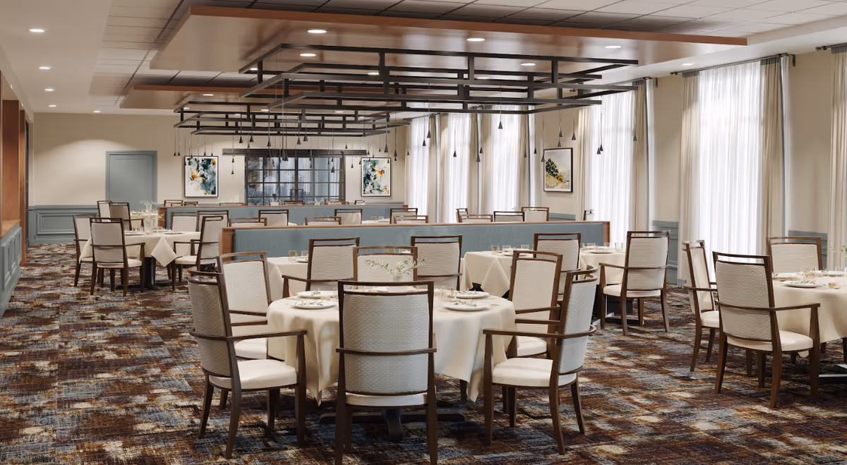 Spacious dining room with round tables set with white tablecloths and upholstered chairs under modern ceiling fixtures and large windows.