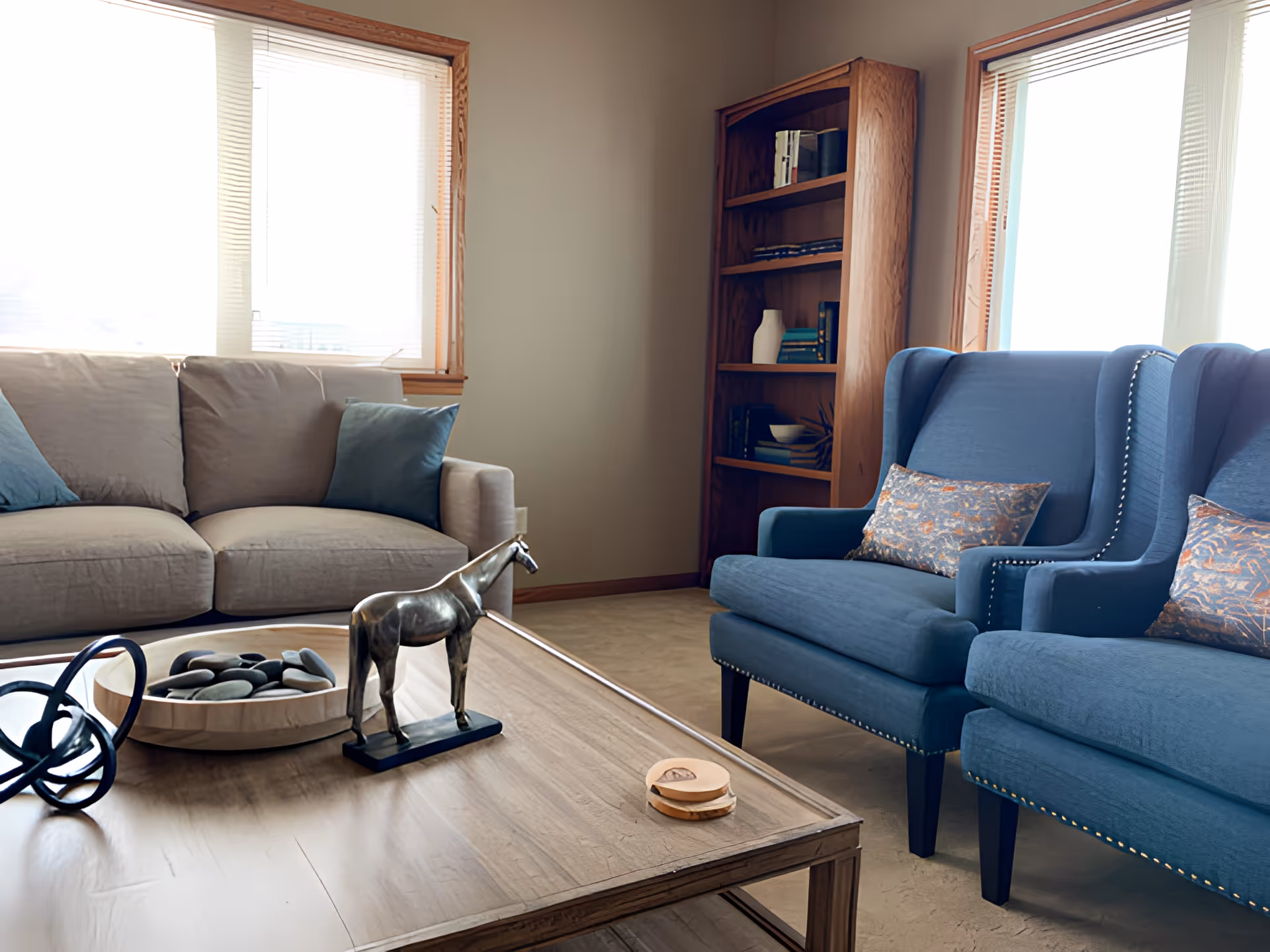 Sunlit living room with a beige sofa, two blue armchairs, a wooden coffee table holding a horse sculpture and decorative bowl, and a bookshelf by the window.