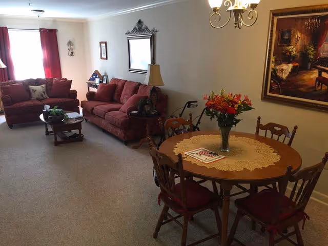 Cozy living room and dining area with red upholstered sofas, a round wooden dining table with a floral centerpiece, wall art, and a coffee table.