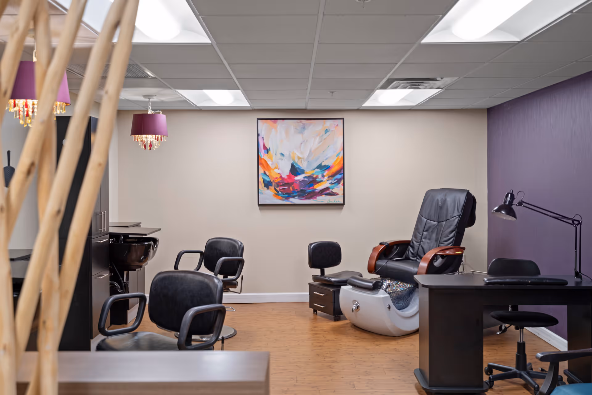 Interior view of a salon area in a senior living facility with black salon chairs, a pedicure chair, a desk with a lamp, and a colorful abstract painting on the wall. The room has a wooden floor and a purple accent wall.