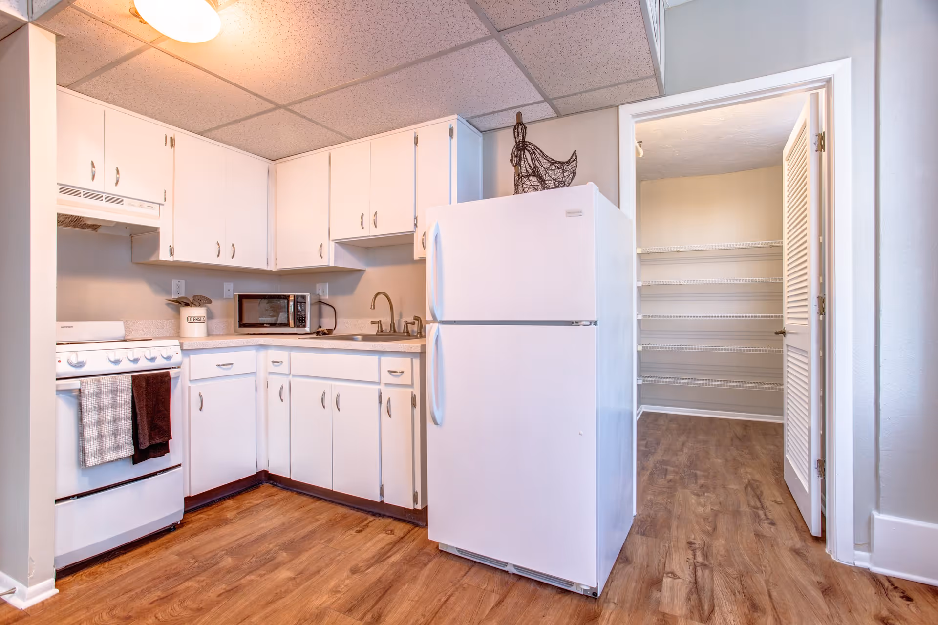 A kitchen interior featuring white cabinets, a white refrigerator, and a stove with a countertop. The space has wooden flooring and a doorway leading to a pantry with shelves.