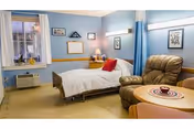 A cozy bedroom in a senior living facility featuring a single bed with white bedding and a red pillow, a comfortable brown recliner chair, a small round table with a decorative centerpiece, a window with blue curtains, framed pictures on the light blue walls, and a nightstand with a lamp and clock.