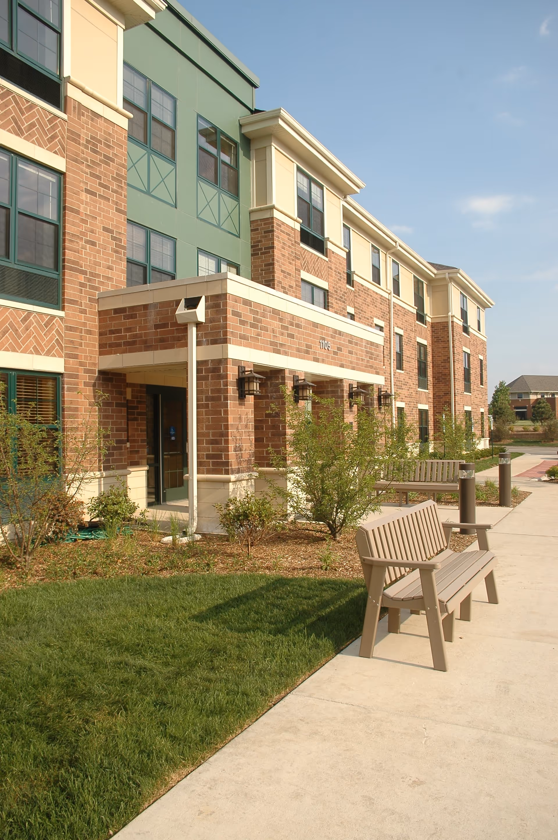 Exterior view of a multi-story brick building with green and beige accents, featuring an entrance with a covered porch. There are benches and small bushes along a concrete walkway in front of the building under a clear sky.