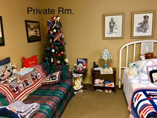 A cozy private room in a senior living facility featuring a green and red patterned couch with patriotic-themed pillows and a quilt, a decorated Christmas tree with American flags, a small side table with a lamp and various items including an Elvis Presley photo, and a white metal bed frame with a colorful quilt and stuffed animals. The walls are beige with framed vintage-style artwork, and the carpet is light brown.