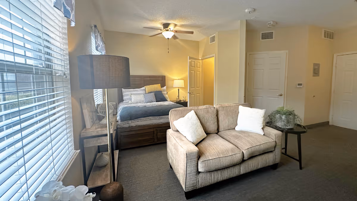 A cozy senior living facility room featuring a bed with multiple pillows, a beige loveseat with two white cushions, a floor lamp, a side table with a plant, and large windows with blinds allowing natural light to fill the space.