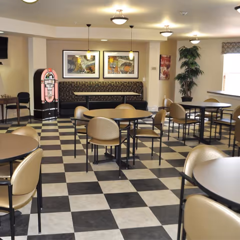 A dining area with multiple round tables and beige chairs on a black and white checkered floor. The room features a cushioned bench along the back wall with two framed pictures above it, a pink vintage jukebox to the left, a potted plant near a window on the right, and ceiling lights providing illumination.