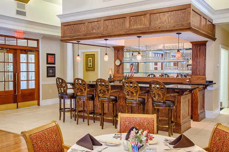 Interior view of a senior living facility dining area featuring a wooden bar with six ornate bar stools, pendant lights hanging above the bar, and a round dining table set with plates, napkins, and a floral centerpiece. The background includes double wooden doors and framed artwork on the walls.
