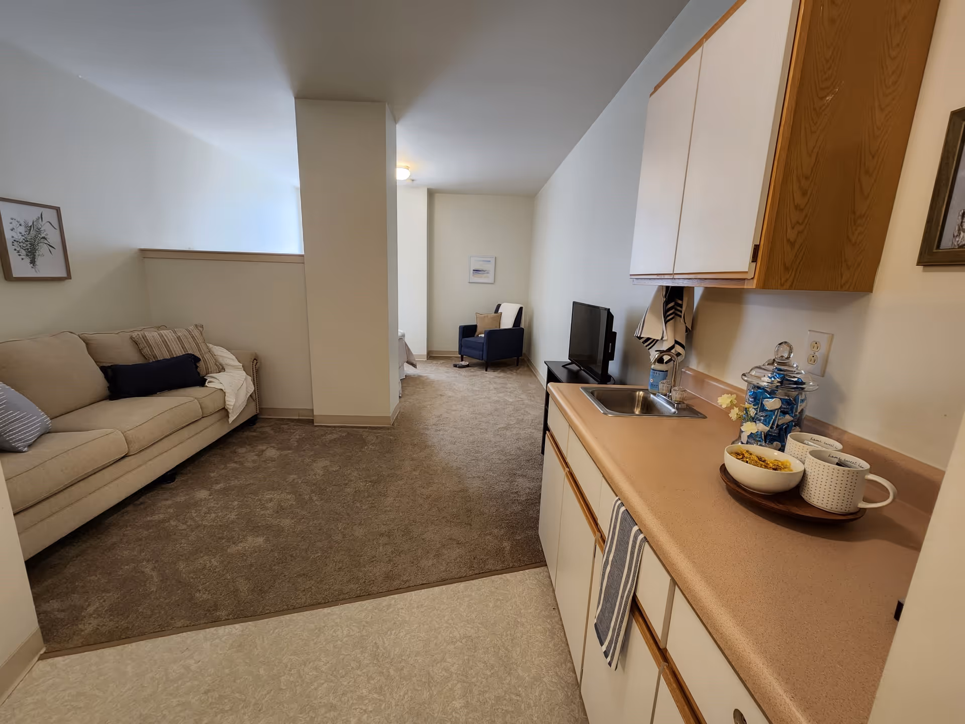Interior view of a senior living facility room showing a small kitchenette with a sink, countertop, and cabinets on the right. On the left, there is a beige sofa with pillows and a blanket. Further back, a blue armchair with a pillow is positioned near a wall with a framed picture. The floor transitions from linoleum in the kitchenette area to carpet in the living and sitting area.