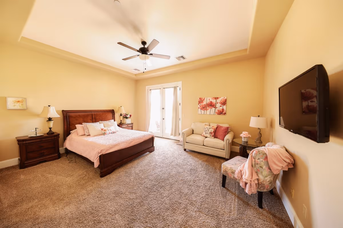 A spacious bedroom with beige walls and carpeted floor. The room features a large wooden bed with pink bedding and multiple pillows, two wooden nightstands with lamps, a small beige loveseat with decorative pillows, a floral upholstered chair with a pink throw blanket, a round side table with a lamp and flowers, a wall-mounted flat-screen TV, and double glass doors with curtains leading outside. A ceiling fan with lights is mounted on the ceiling.