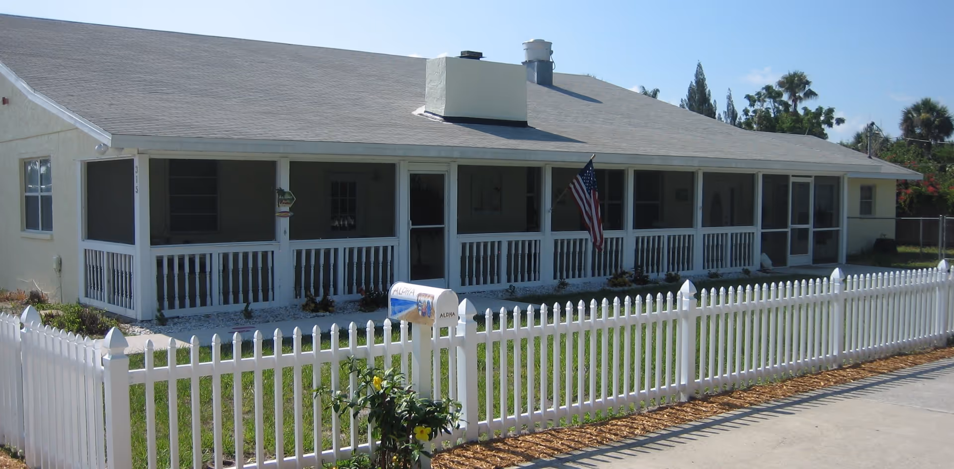 Single-story residential building with a light-colored exterior, a screened front porch, an American flag hanging near the entrance, and a white picket fence enclosing a small grassy yard with a mailbox.