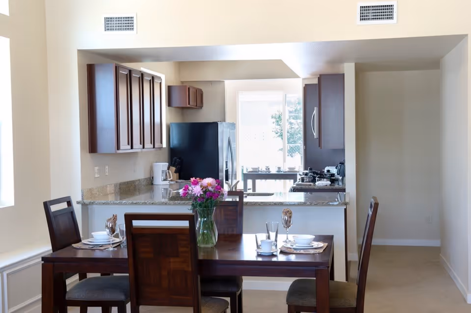Dining table set with dishes and a vase of flowers in an open-plan dining area facing a kitchen with granite counters and a stainless refrigerator.
