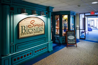 Interior hallway of a senior living facility with teal-colored wall paneling and a large wooden sign that reads 'Simple Blessings Country Store.' Next to the sign is a glass display case and a chalkboard sign advertising store hours. The hallway extends toward a glass door entrance with natural light coming through.