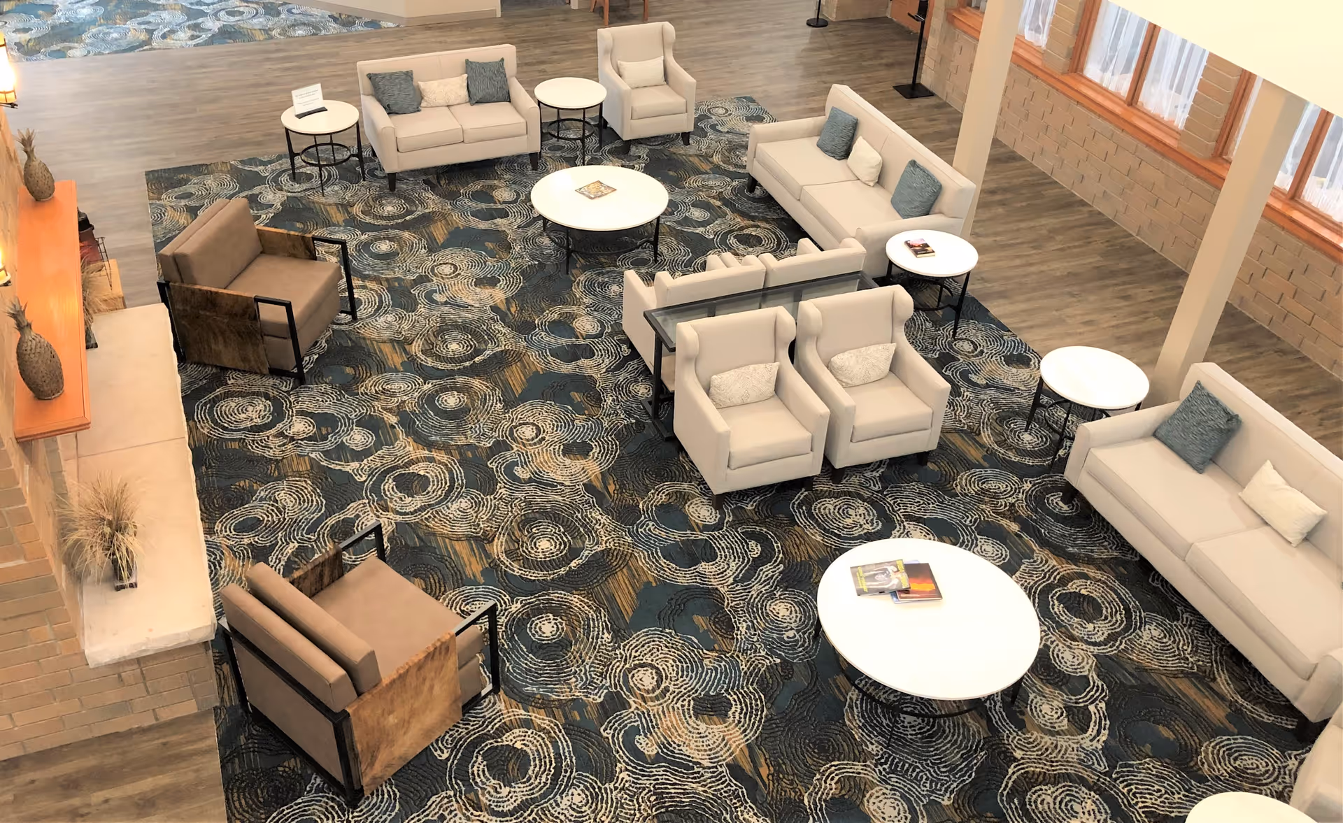 A spacious living room area with multiple beige sofas and armchairs arranged around several round white coffee tables on a patterned blue and beige carpet. Large windows with sheer curtains allow natural light to fill the room. The walls are light-colored with brick accents and wooden trim.