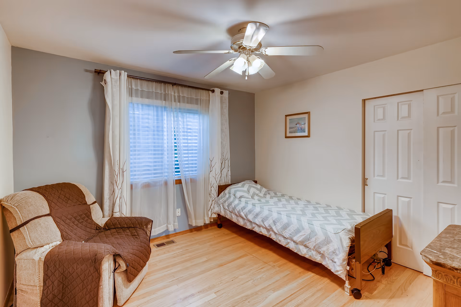 A simple bedroom with a single bed covered in a gray and white chevron patterned blanket, a beige armchair with a brown quilted cover, a window with sheer white curtains, a ceiling fan with lights, a framed picture on the wall, and a wooden floor.