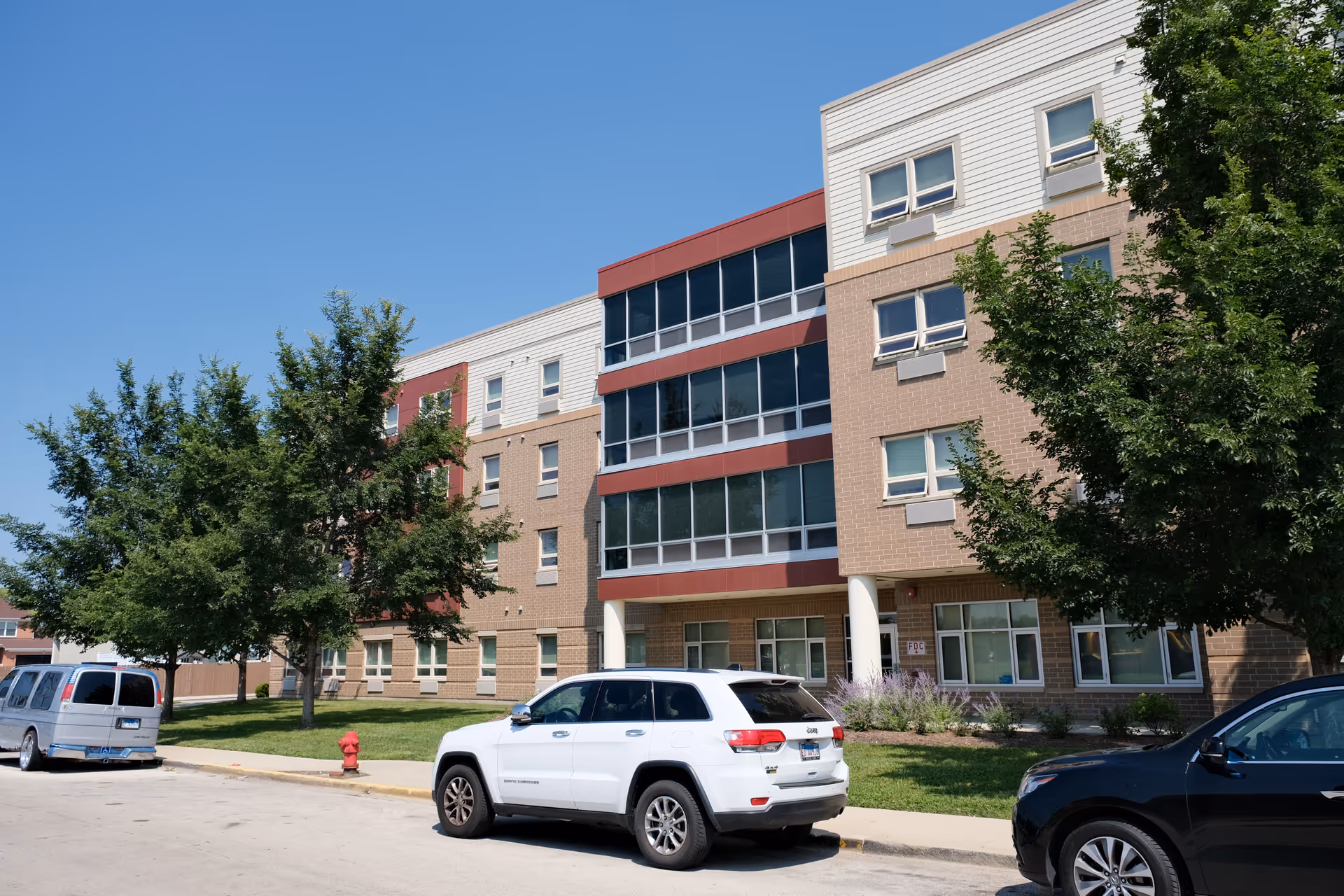 Front exterior of a multi-story senior living building with parked cars, trees, and a clear blue sky.