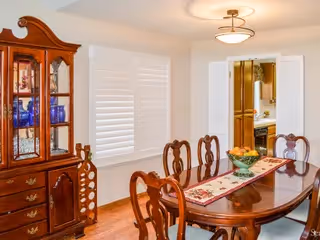 A traditional dining room with a polished wooden dining table and six matching chairs. The table is decorated with a floral table runner and a bowl of fruit. To the left, there is a wooden china cabinet displaying glassware. The room has white walls, a window with white shutters, and a ceiling light fixture. A doorway leads to another room, possibly a kitchen.