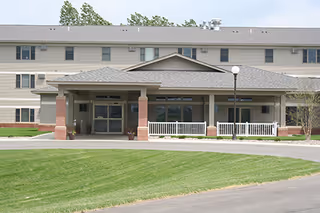 Front exterior view of Shepherd of Grace Senior Community building with a covered entrance supported by brick columns, white railings, and a well-maintained lawn in front.