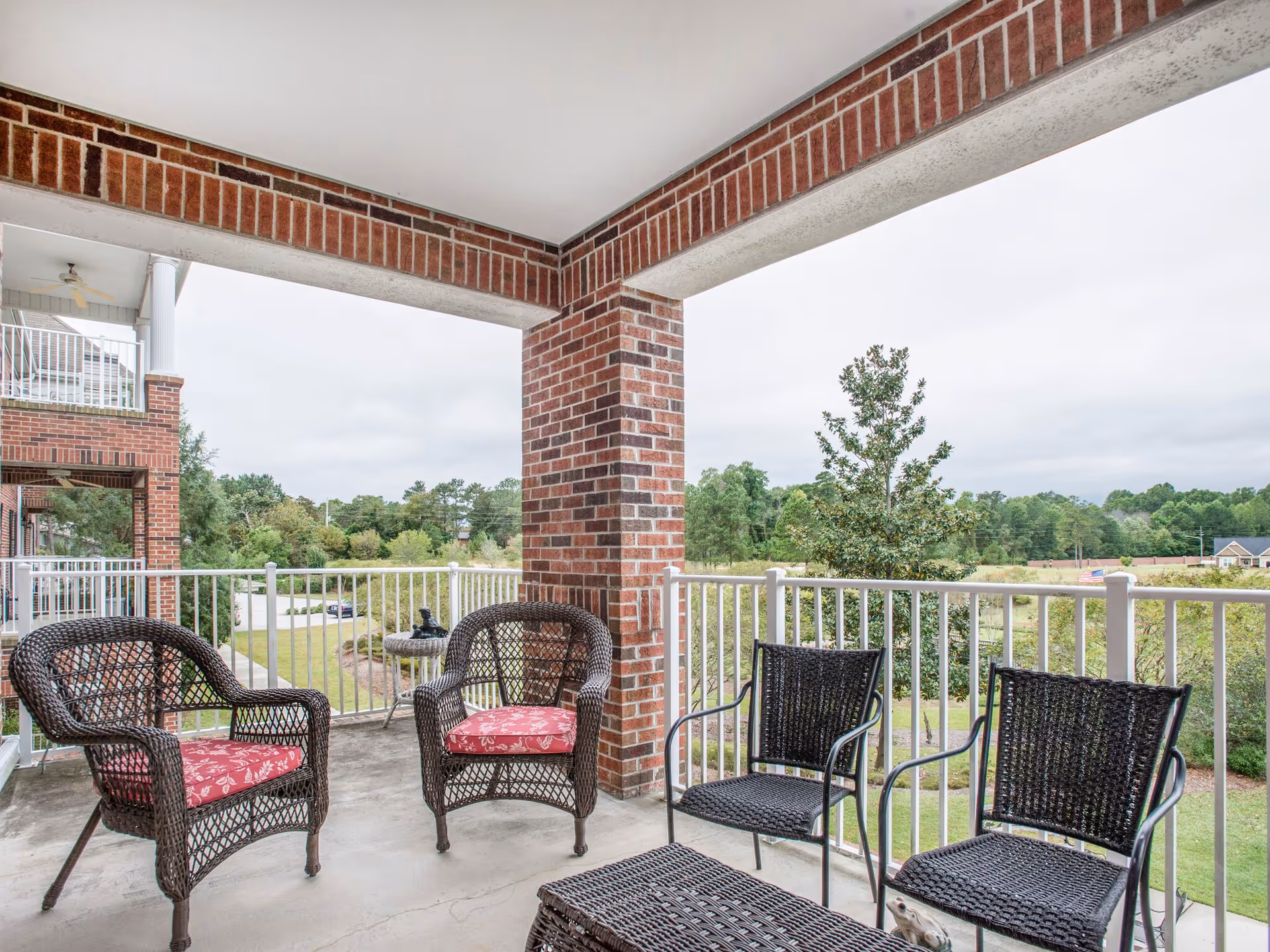 Covered outdoor patio area with brick pillars and white railing, furnished with two wicker chairs with red cushions, two black woven chairs, and a matching black woven coffee table. The patio overlooks a green landscape with trees and a cloudy sky.