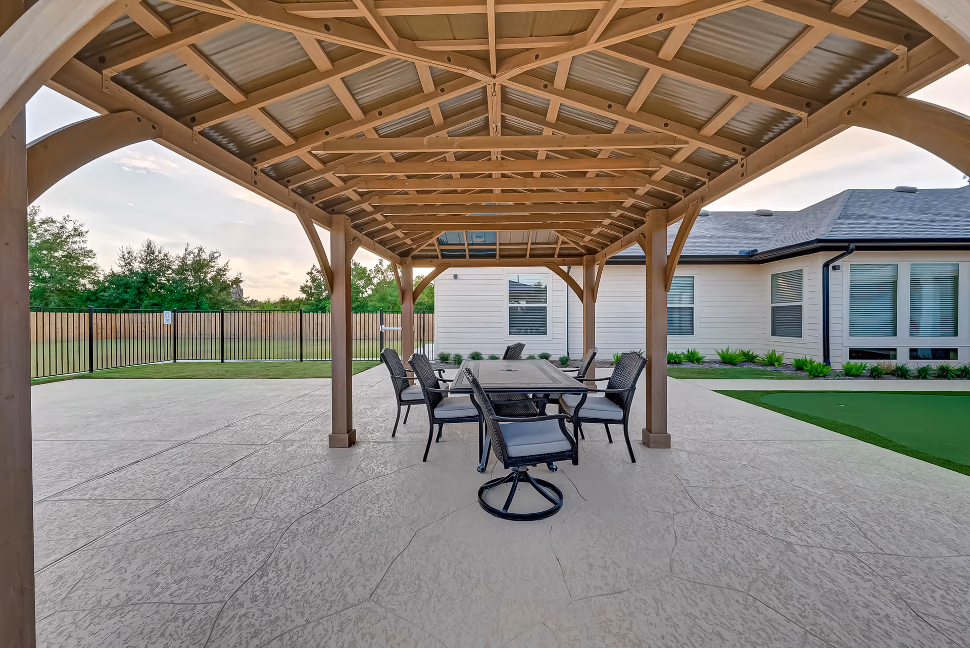 Outdoor covered patio area with a wooden pergola roof, featuring a rectangular table surrounded by six cushioned chairs on a concrete surface. In the background, there is a fenced grassy area and a single-story building with multiple windows and landscaping.