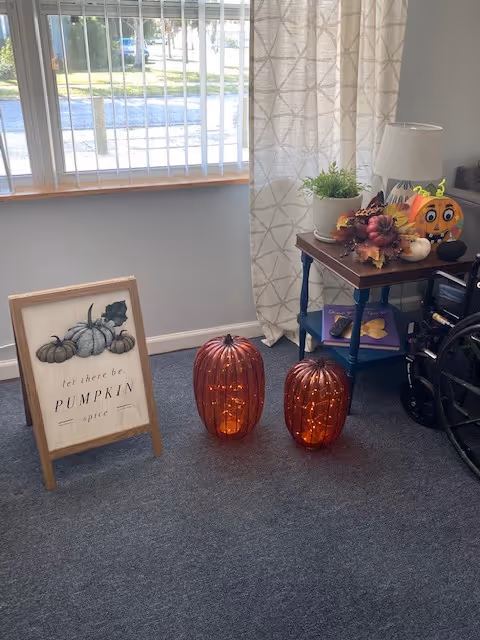 Corner of an interior room with fall decorations including a framed pumpkin sign, two lit decorative pumpkins on the floor, a side table with a lamp and small pumpkins, and a wheelchair.