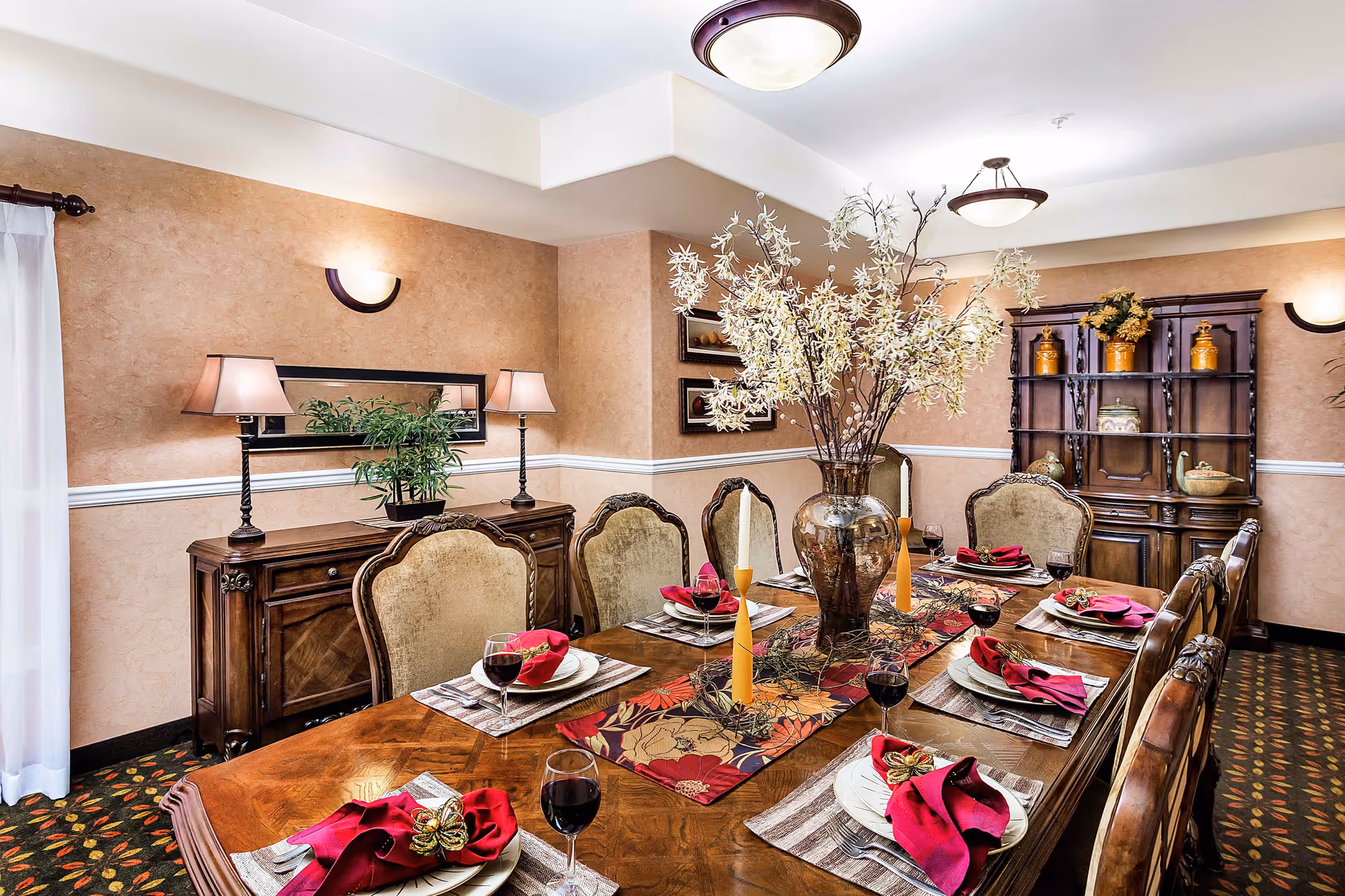 A formal dining room with a long wooden table set for six people. Each place setting includes a white plate, a red folded napkin with a decorative napkin ring, silverware, and a glass of red wine. The table has a floral centerpiece with tall candles. The room features beige walls, a wooden sideboard with two lamps and a plant, and a wooden cabinet with decorative items. The carpet has a floral pattern, and the room is lit by ceiling lights and wall sconces.