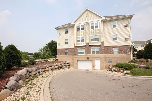 Exterior view of a three-story senior apartment building with beige siding and brick accents. The building has multiple windows and a garage door at the ground level. The surrounding area includes a curved driveway, landscaped rock borders, and green shrubs.