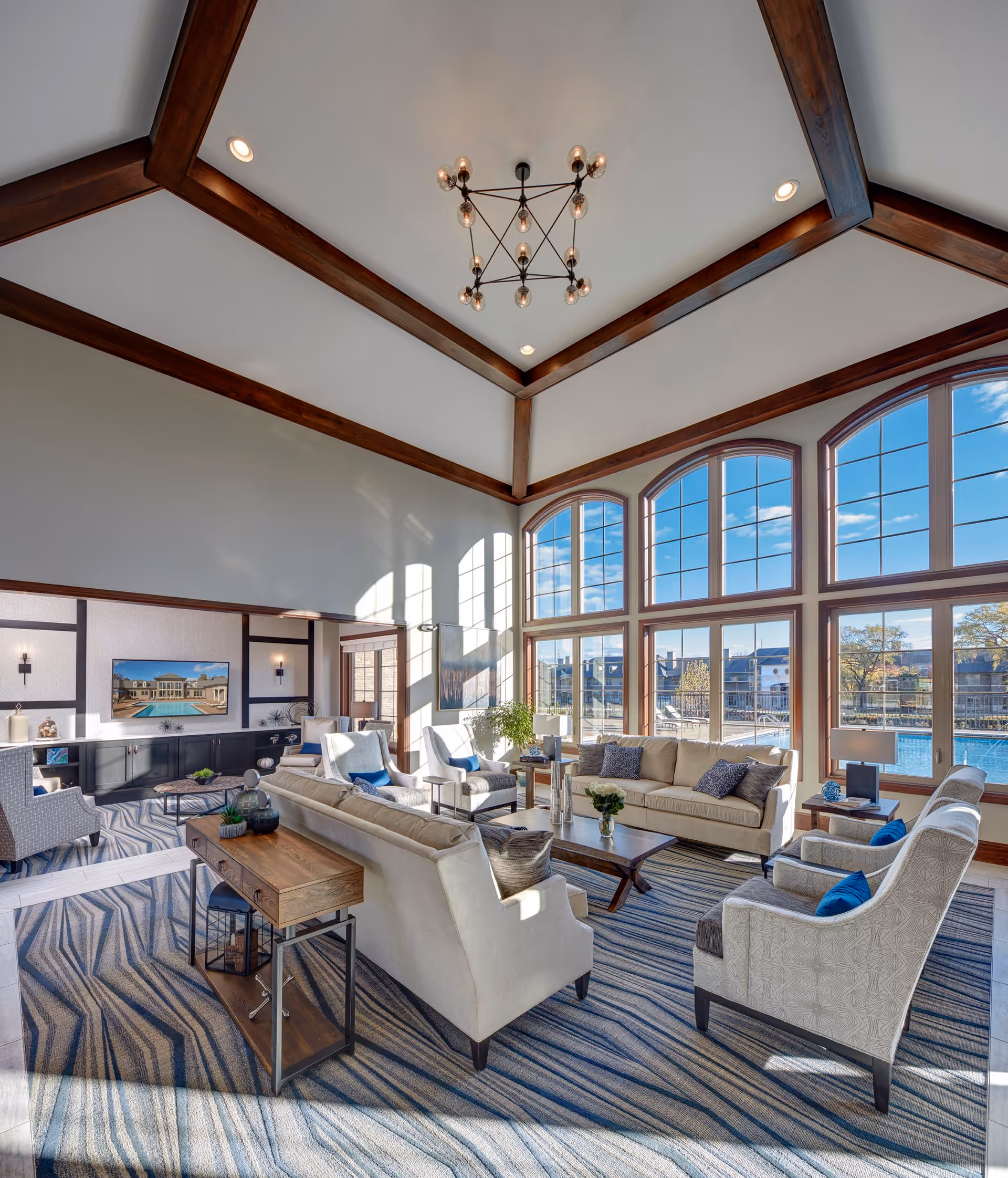 A spacious and bright living room with large arched windows letting in natural light. The room features beige sofas, patterned armchairs with blue cushions, a wooden coffee table, and a side table with decorative items. The ceiling has wooden beams and a modern chandelier. Outside the windows, a pool and neighboring buildings are visible.