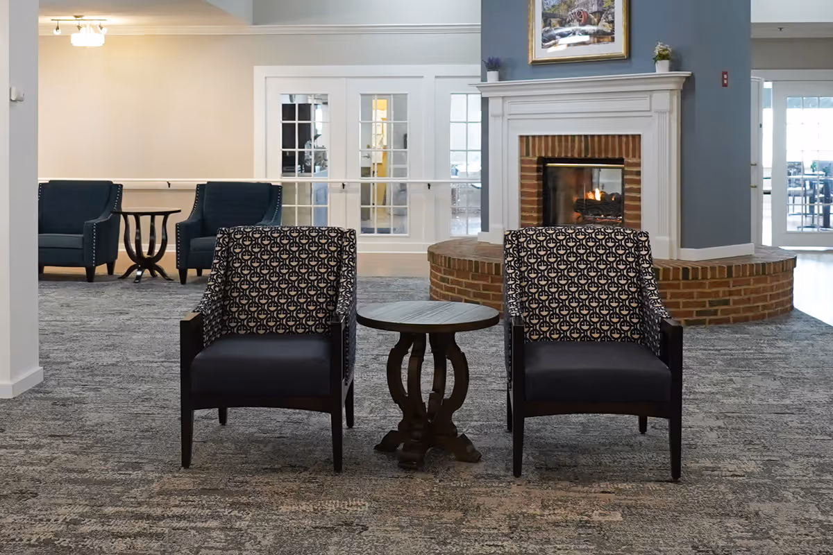 A cozy seating area in a senior living facility featuring two patterned armchairs with a small round wooden table between them. In the background, there is a brick fireplace with a fire burning, a painting above it, and additional seating with two blue armchairs and a small table. The room has carpeted flooring and glass-paneled double doors.