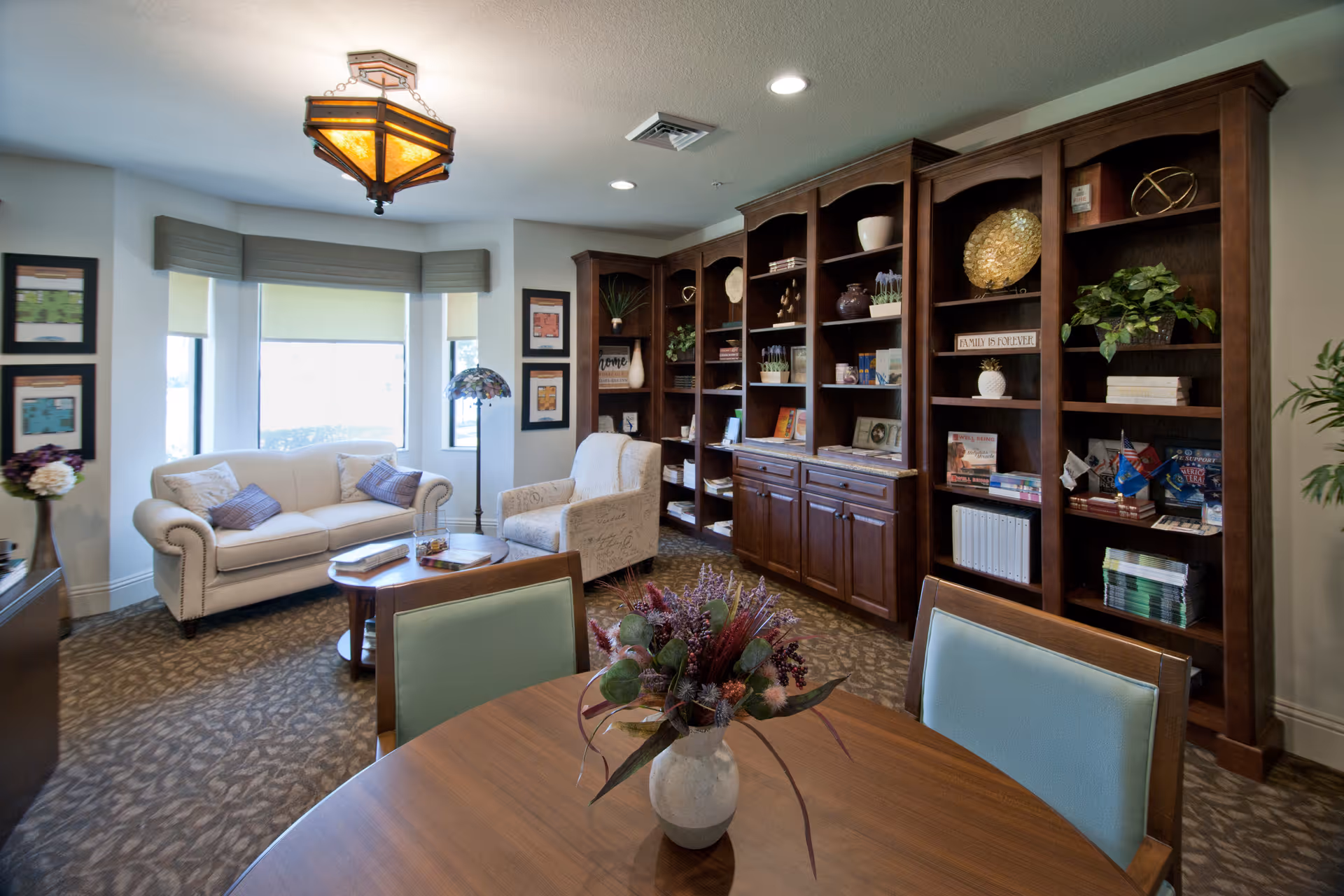 A cozy living room area with a white sofa, an armchair, a wooden coffee table, and a round wooden dining table with chairs. The room features a large wooden bookshelf filled with books and decorative items, a window with blinds, framed pictures on the walls, a floor lamp, and a ceiling light fixture.